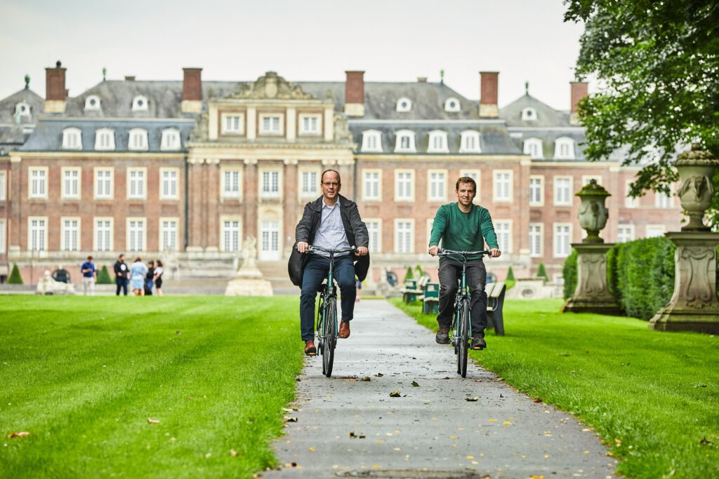 Zu sehen sind zwei Radfahrer vor der Kulisse des Schlosses in Münster.